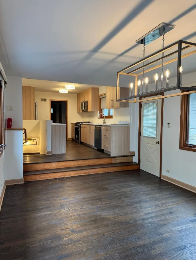 Dining area with dark-stained hardwood flooring and a black linear chandelier, stepping up to a kitchen with light wood cabinets