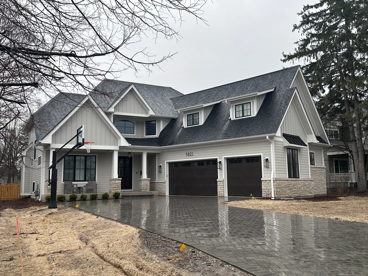 Two-story home with white board-and-batten and lap siding, a stone veneer base, dark shingled roof with gables and dormers, two dark garage doors and a paver driveway