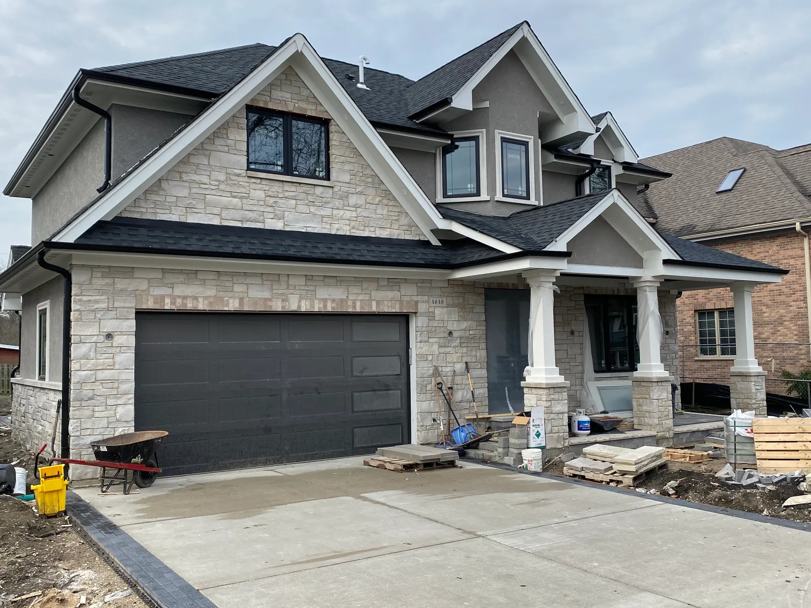 Two-story home with a stone veneer front and gray stucco upper story, a dark shingled roof with gables and dormers, a dark two-car garage door, a covered front porch on tapered columns and a newly poured concrete driveway edged with pavers