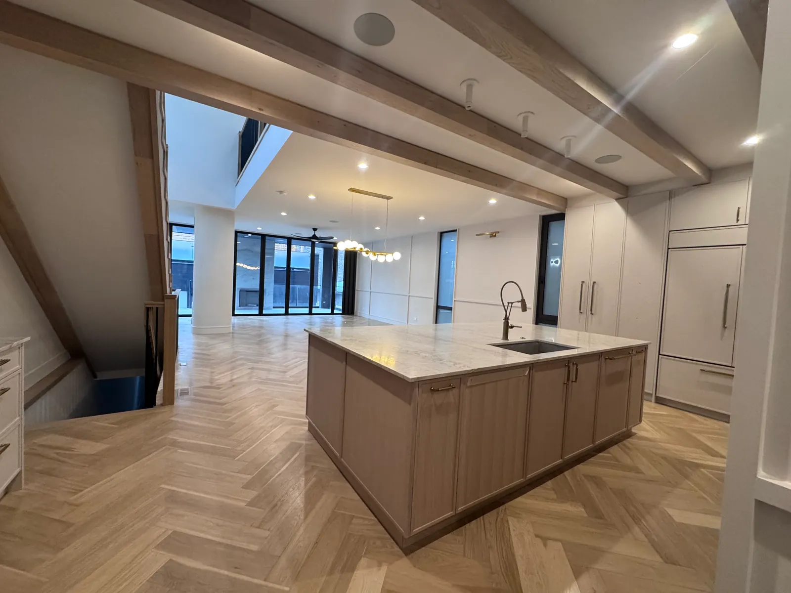 Open-plan kitchen with a large white oak island, a marble-look countertop and undermount sink, exposed white oak ceiling beams and herringbone wood flooring running through to a living area with floor-to-ceiling black-framed windows
