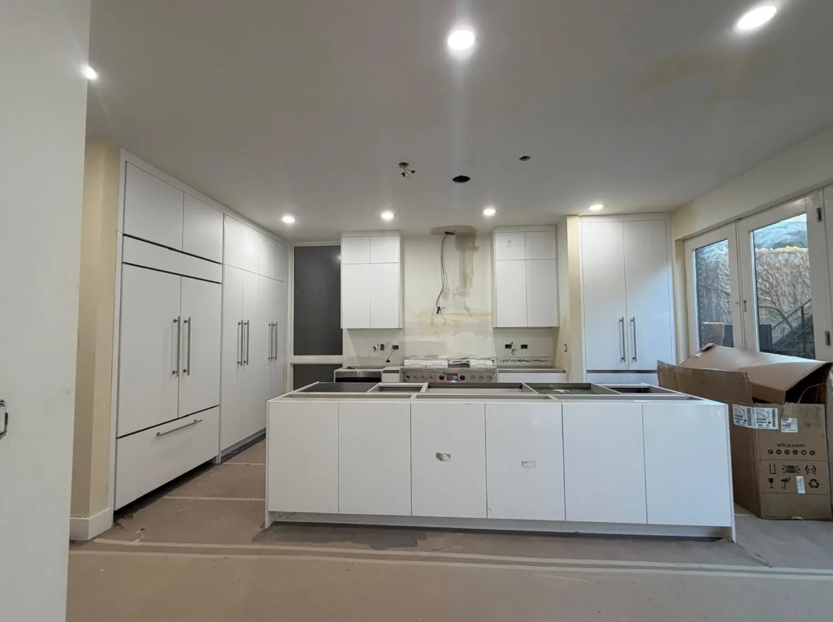 Kitchen mid-installation with white slab-front cabinets, a tall panel-front refrigerator and island cabinets set in place, floor still covered and countertops not yet fitted