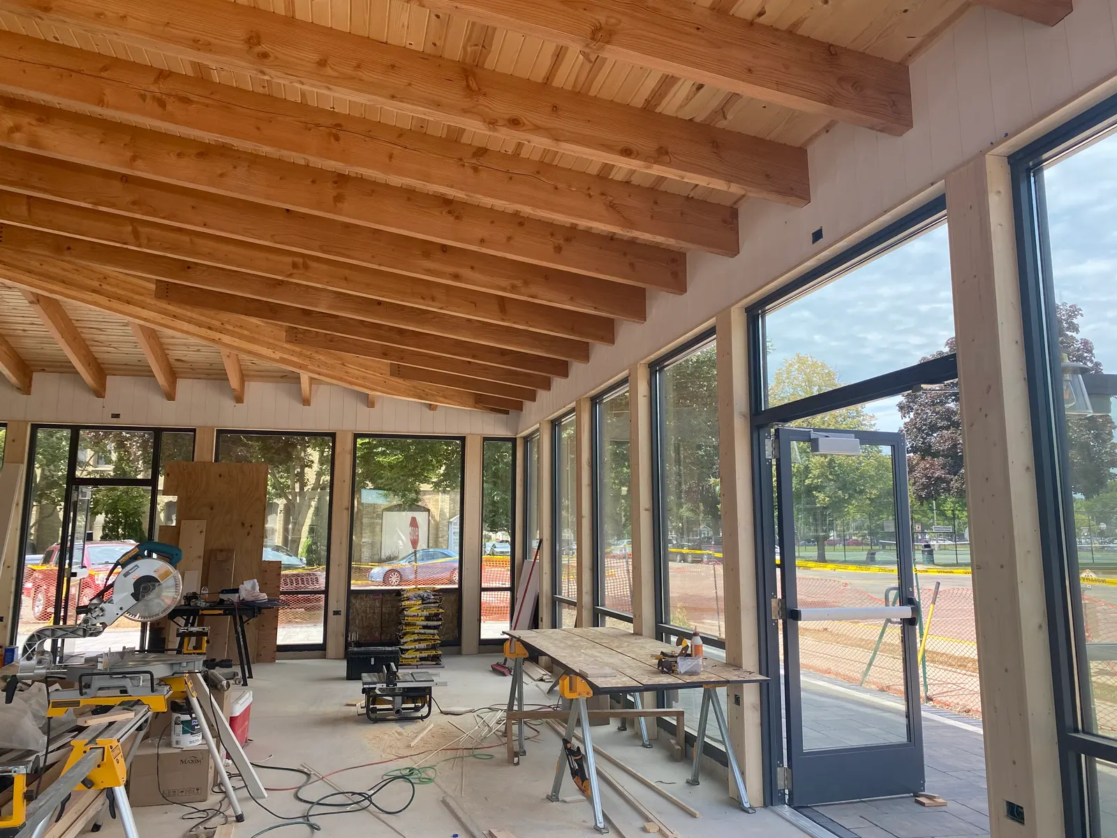 Interior under construction with an exposed timber beam ceiling and wood plank decking above a wall of black-framed windows and glass doors, with a miter saw and sawhorses set up on the bare concrete floor