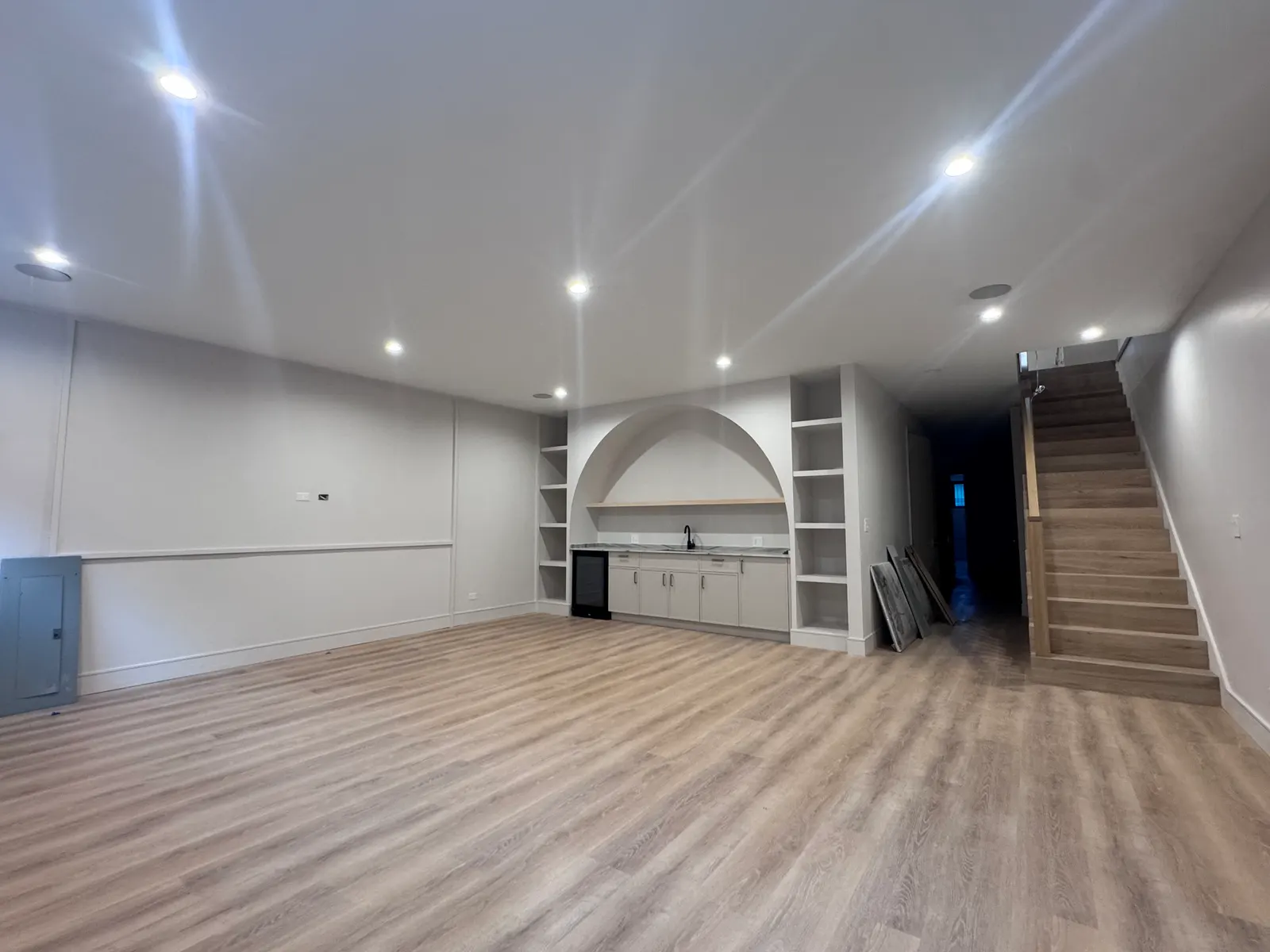 Open finished room with light wood-look plank flooring and recessed ceiling lights, a wet bar of white cabinets with a sink and a beverage refrigerator set into an arched alcove flanked by built-in shelving, and a wood staircase at the right
